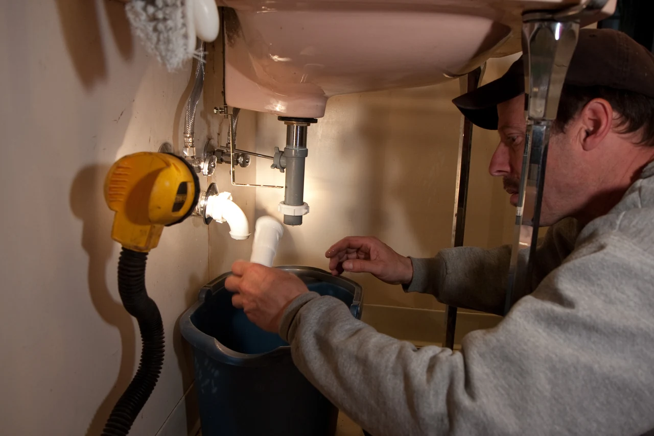 A plumber repairing the drain beneath a sink, with a work light and a bucket under the open pipes.